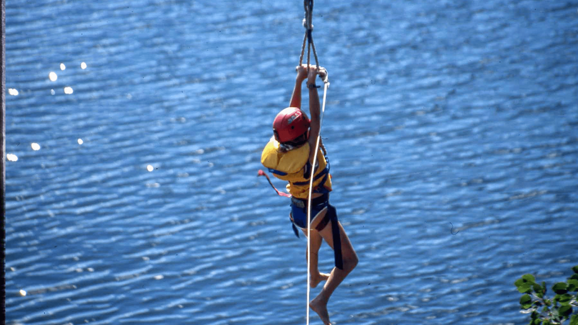 A child climbing a tall rope over a blue lake.