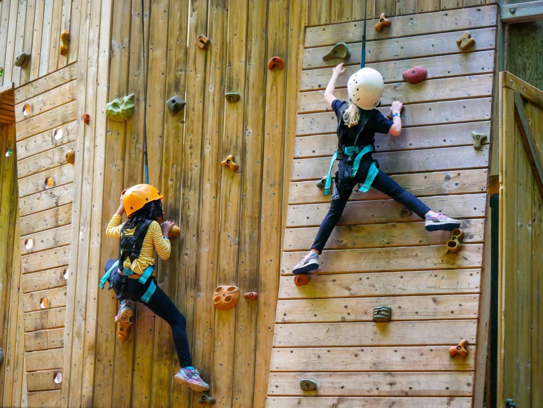 Two children climbing a wooden rock wall at summer camp.