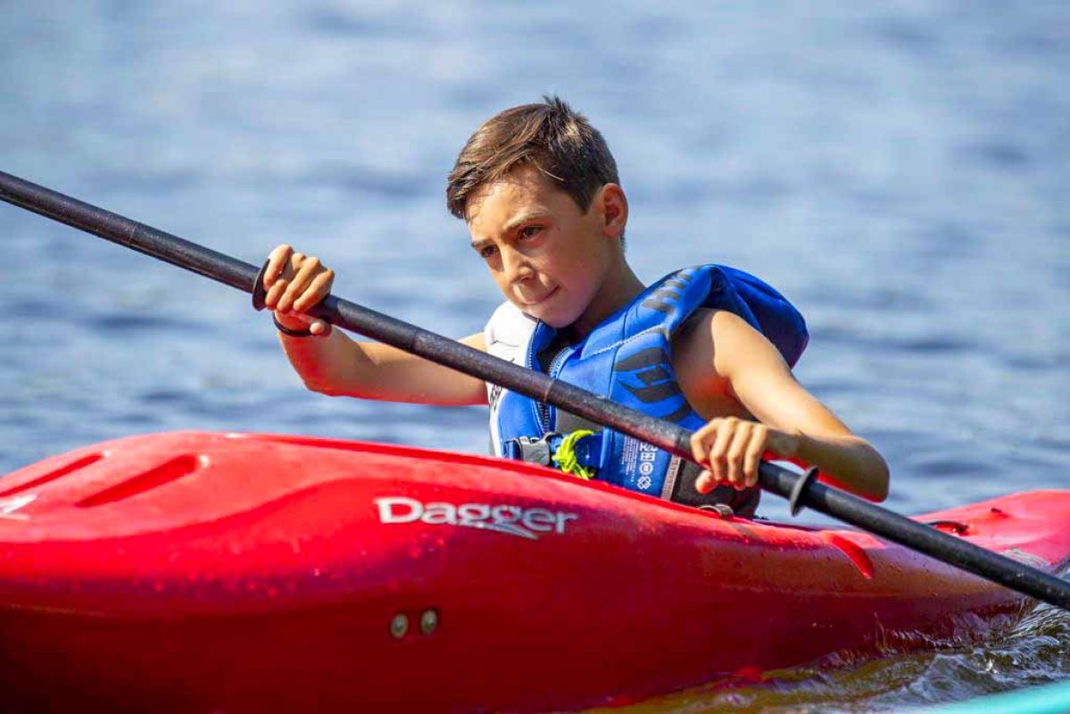 A young boy paddling a red kayak on a lake.